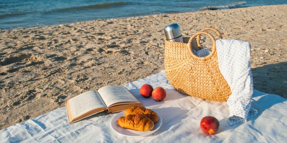 Summer picnic on the beach by the sea at sunrise. There is a white picnic blanket with a beach bag, a book, two croissants, and three peaches on the picnic blanket.
