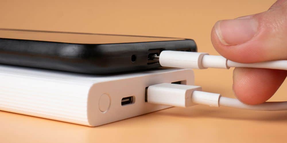 A closeup image of a smartphone placed on top of a portable power bank, with a person's thumb and forefinger plugging a white USB cable into the phone.
