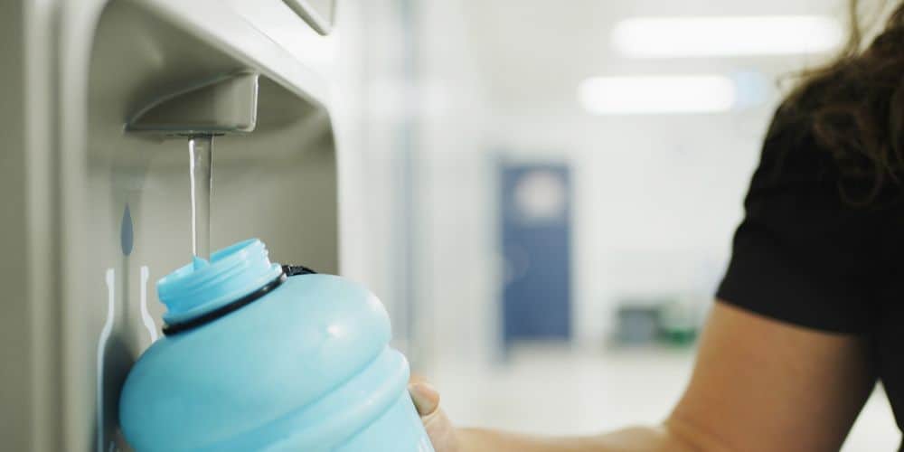 A person is refilling a blue sports water bottle from a water dispenser in a blurry indoor setting.