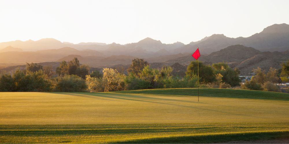 Golf green at dusk with a red flag in the hole, set against a backdrop of mountains and desert terrain.