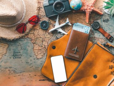 Travel essentials laid out on a world map, including a straw hat, red sunglasses, a vintage camera, compass, starfish decoration, airplane model, passport holder, wristwatch, and a smartphone.