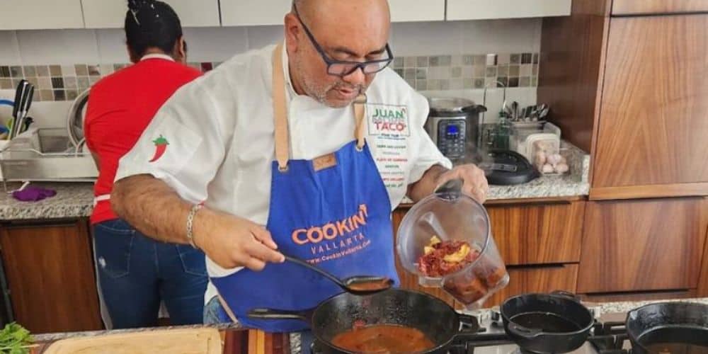A man in a blue apron labeled "COOKIN' VALLARTA" and a white chef's coat is attentively cooking, tipping a clear container with food into a sizzling pan during a cooking class in Cabo.