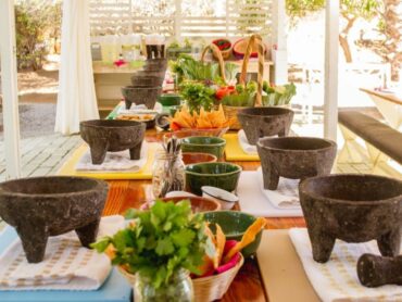 A cooking class setup outdoors with multiple molcajetes (traditional Mexican stone mortar and pestle) on a long table, ready for use. Fresh ingredients are arranged around the table, including a variety of vegetables and tortilla chips in baskets.