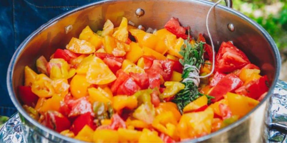 A closeup of a stainless steel pot filled with a vibrant mix of chopped tomatoes in hues of yellow, orange, and red, with a bundle of fresh herbs including what appears to be thyme.