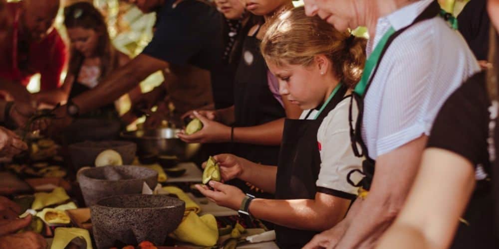 A cooking class in progress with participants of all ages around a table, preparing ingredients. At the center is a young girl intently holding an avocado.