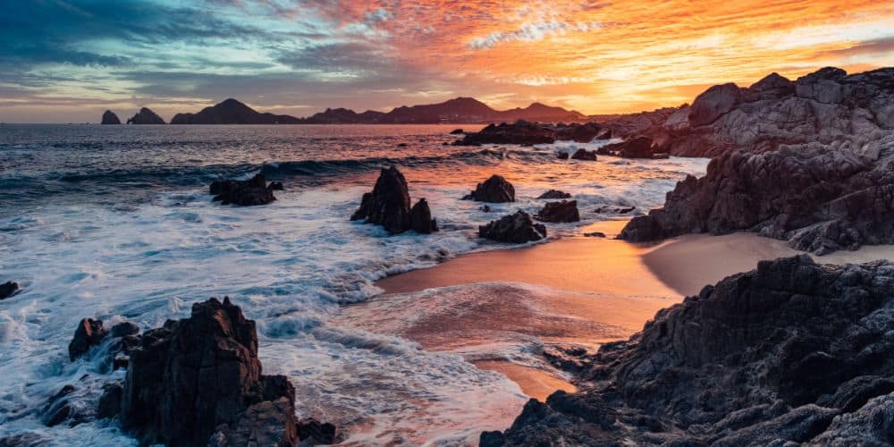 A dramatic sunset over a rugged beach with fiery skies reflecting on the water's surface. Jagged rocks and boulders punctuate the shoreline, as frothy waves wash onto the sandy coves.